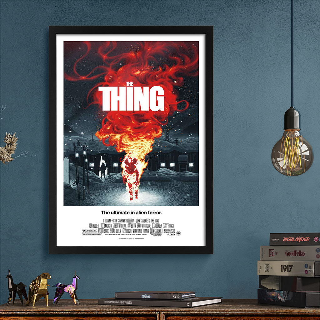 Movie poster for 'The Thing' framed on a blue wall with books and decorative items below.